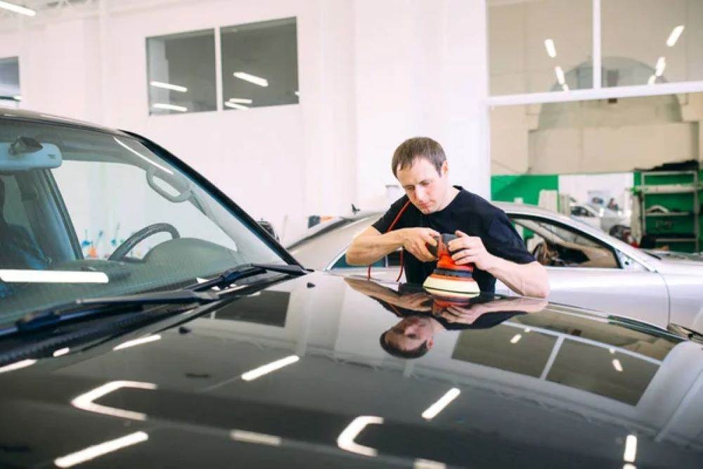 Man utilizes advanced car cleaning techniques with a buffer on a vehicle's hood in a bright, modern garage
